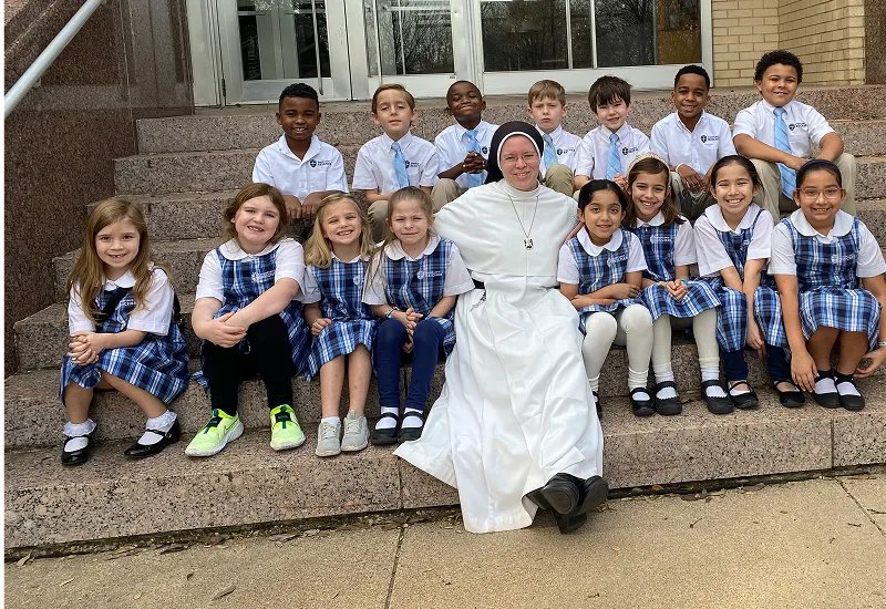 A of diverse students in blue and white uniforms sits on the front steps of a Catholic school surrounding their Catholic elementary school teacher, a religious sister who has been using a Catholic virtue education program for spiritual growth by Sister John Dominic.
