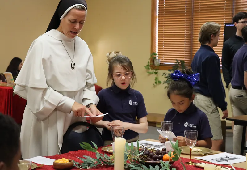 sister-with-kids-at-festive-table_compressed A Dominican Sister in traditional habit assists two young students wearing school uniforms with a lesson at a table adorned with candles, fruit, and educational papers. The setting includes a red tablecloth, greenery, and a warm indoor environment with wooden blinds and other students in the background, highlighting a Catholic faith-based educational experience.